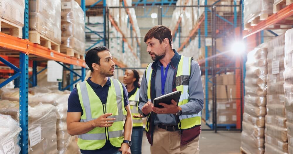 Two men in neon vests walk through a warehouse.